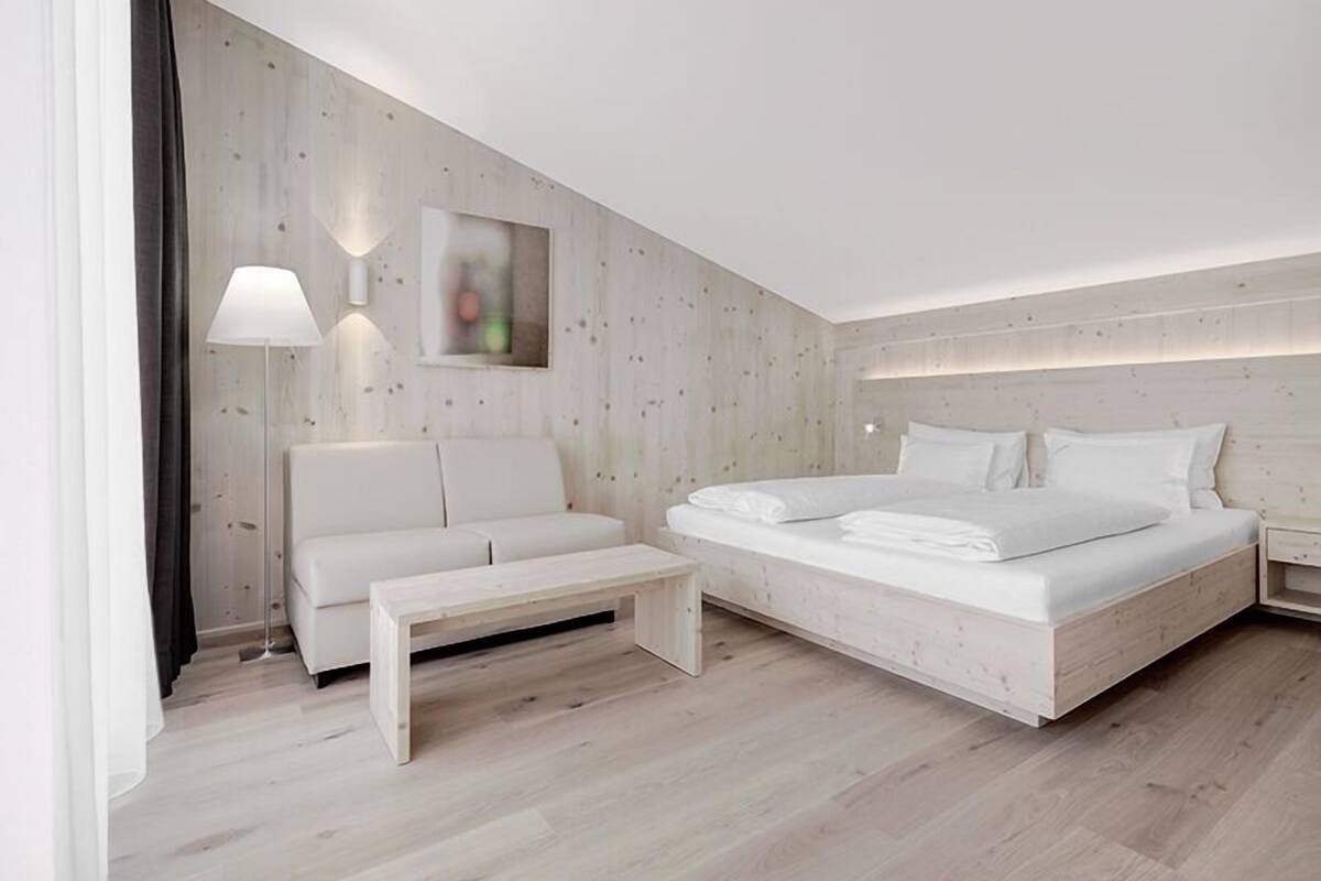 A modern hotel room with a wooden wall, a white bed, a white sofa, a small wooden table, and a floor lamp.
