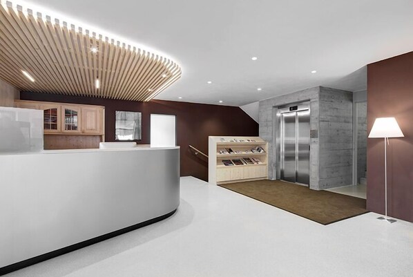 A modern reception area with a curved white reception desk, a wooden slatted ceiling, a magazine rack, and a marble elevator. - Hotel Gardenazza (Badia)