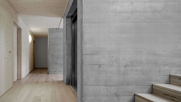 Un pasillo con piso de madera, paredes de concreto y una escalera con escalones de madera.