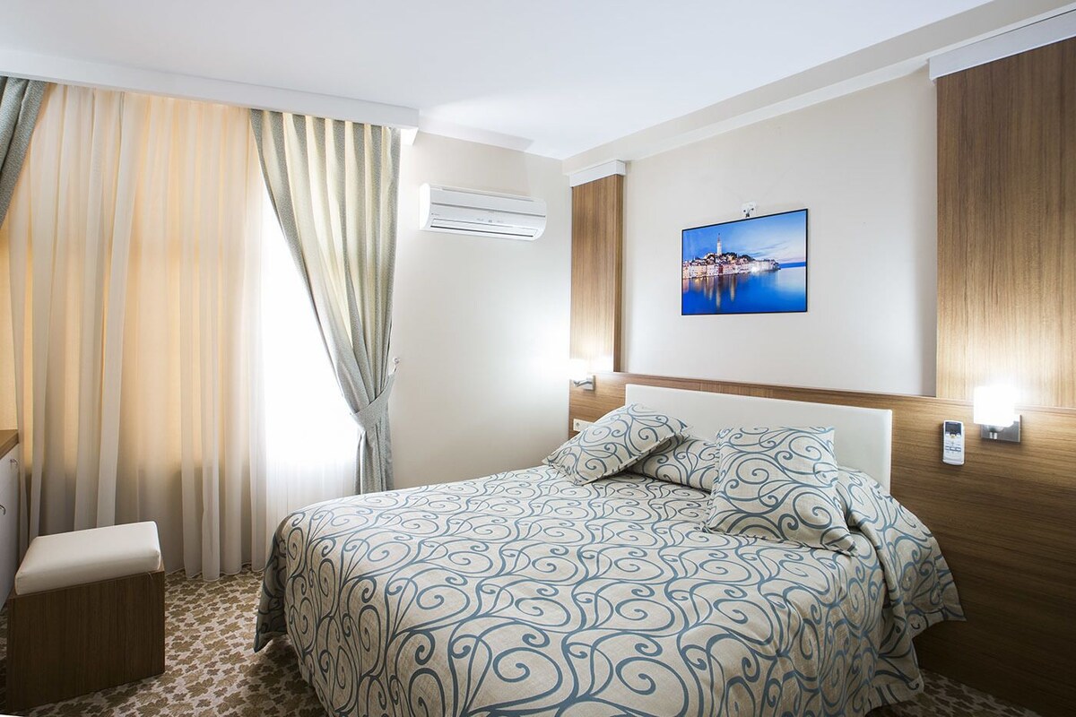 Ein Hotelzimmer mit Bett, Nachttisch, Klimaanlage, Wandfernseher und einem Fenster mit Vorhängen.