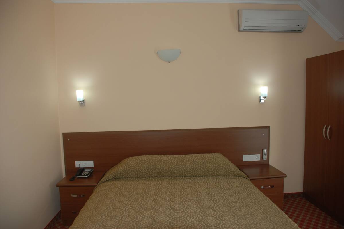 A hotel room with a bed, bedside tables, a wall-mounted air conditioner, and two wall sconces.