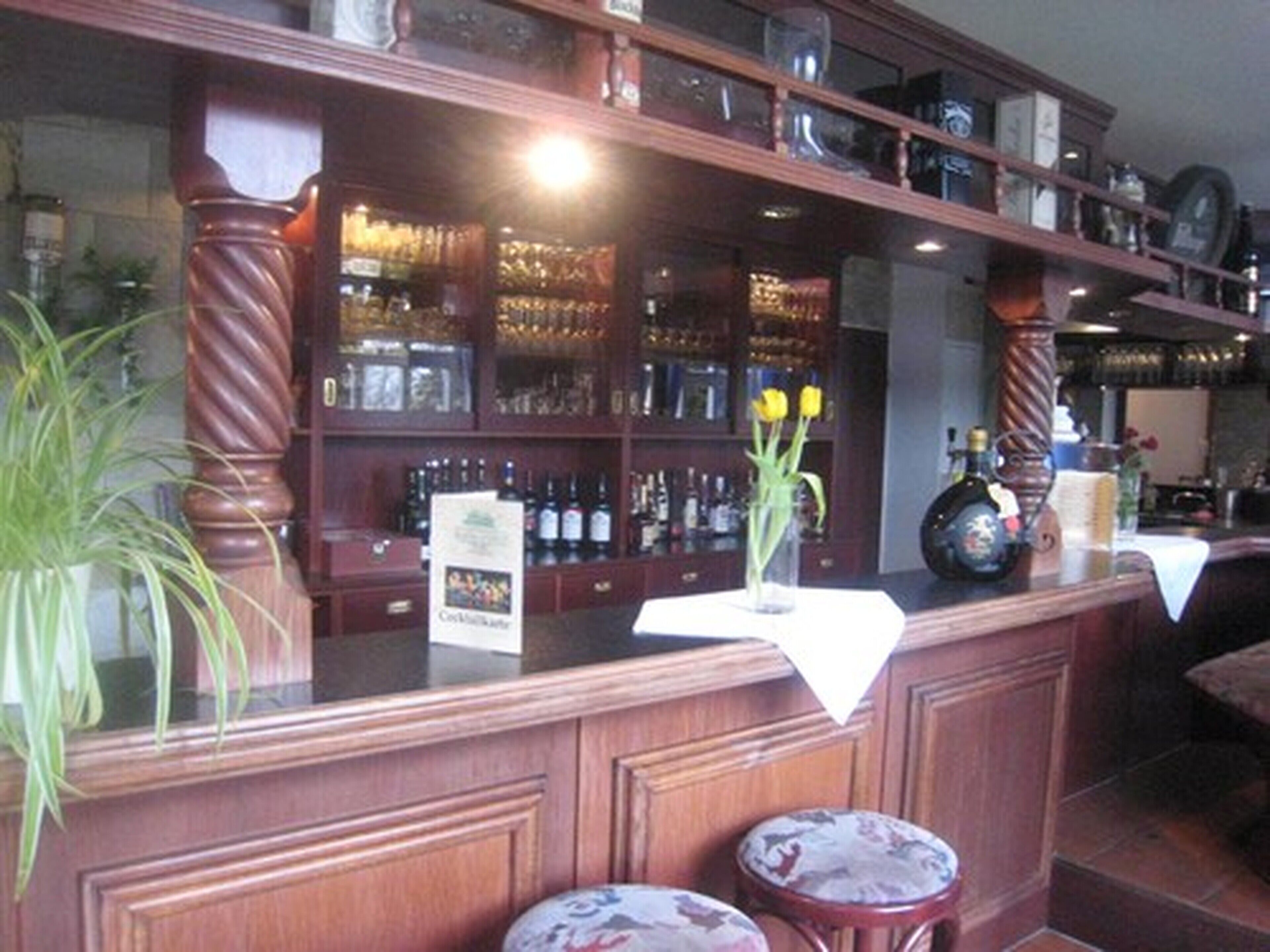 A bar area with wooden shelves stocked with bottles, a vase with flowers, and two stools.