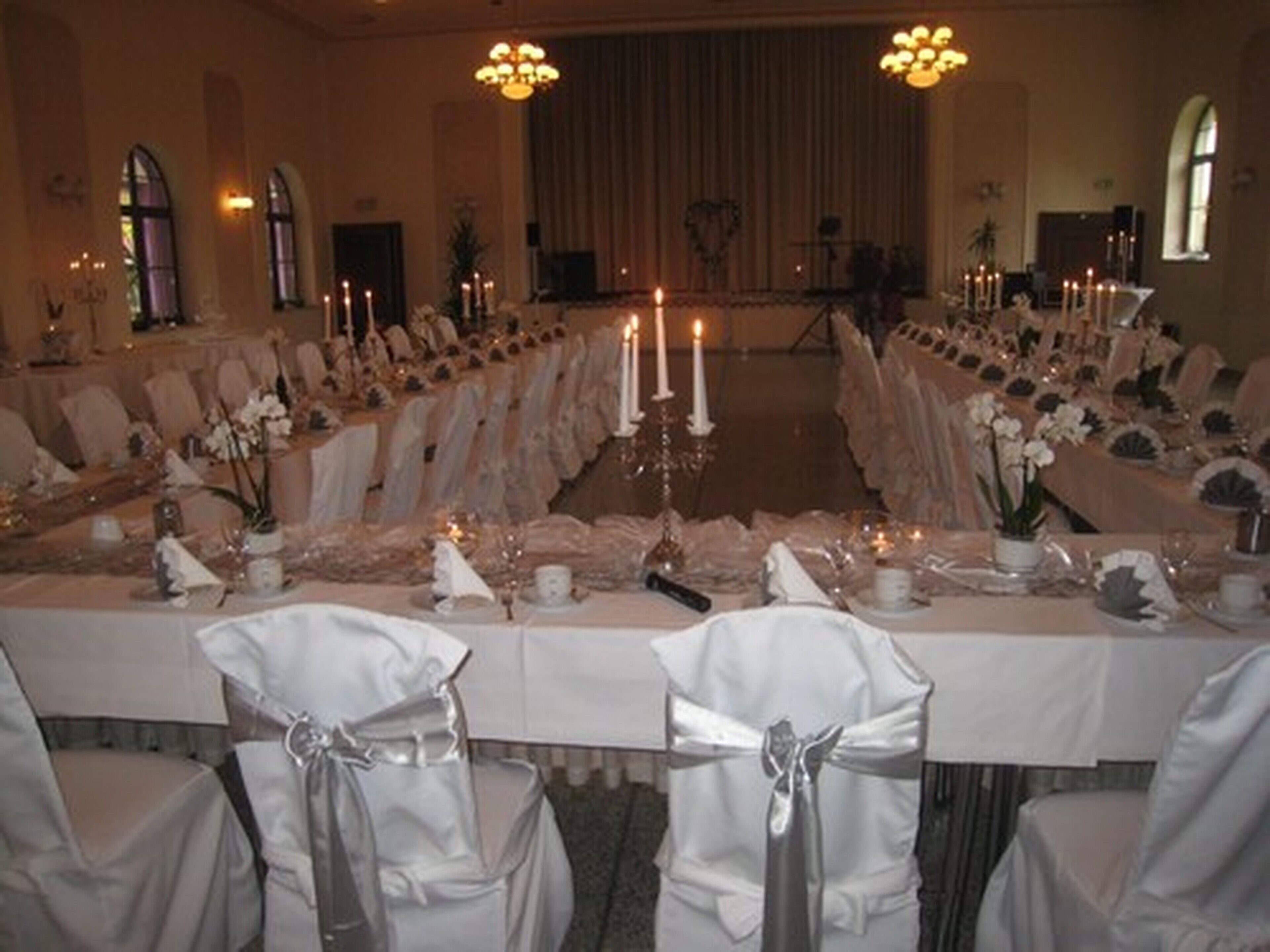 Un salón de banquetes con largas mesas dispuestas para un evento formal, con manteles blancos, velas y centros de mesa florales.