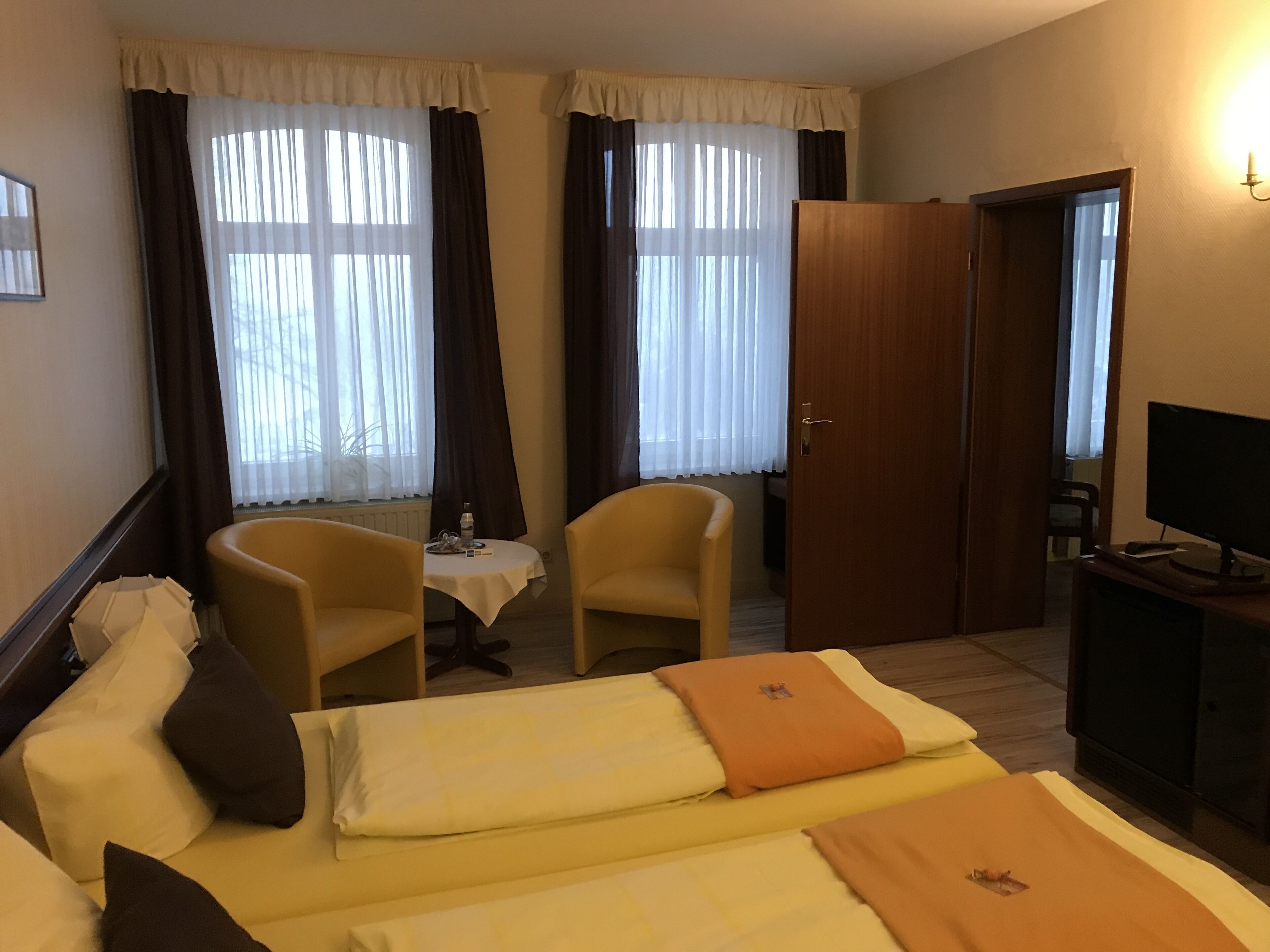 A hotel room with two single beds, a desk with a chair, a television, and two windows with curtains.
