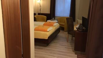 Ein Hotelzimmer mit Holzboden, zwei Betten mit orangefarbener Bettwäsche, einem Schreibtisch mit Computer, einem Sessel und einem Fenster mit Vorhängen.