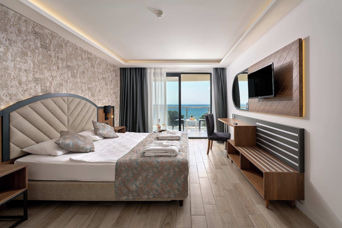 A modern hotel room with a large bed, a TV mounted on the wall, a balcony with a view, and a desk area.