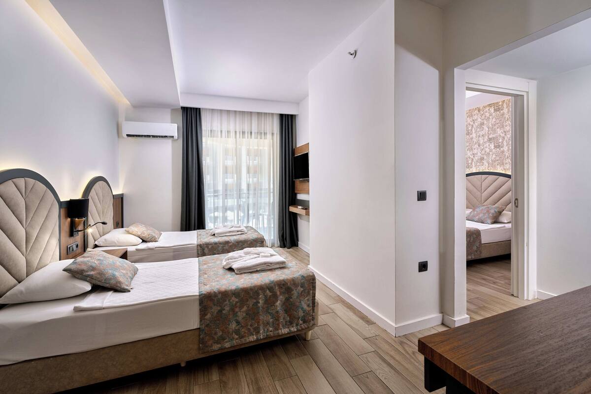 A modern hotel room with two beds, a wooden floor, a large window with curtains, and a view of the city.