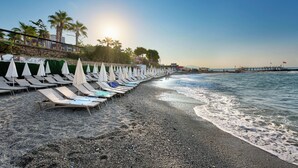 A beach with rows of sun loungers and umbrellas, a stone wall, palm trees, and a pier in the background. - Grand Uysal Beach&Spa Hotel - All inclusive (Alanya)