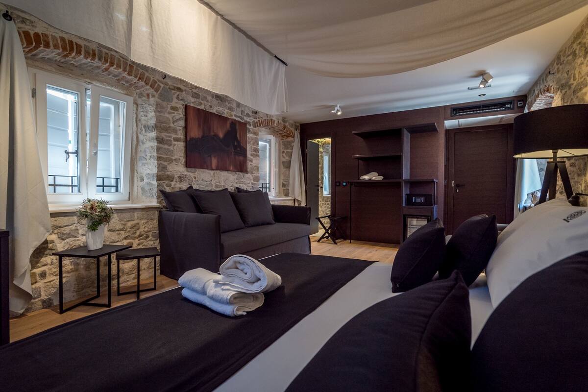 A modern room with a stone wall, a sofa, a bed with rolled towels, a small table with a vase, and a large window.