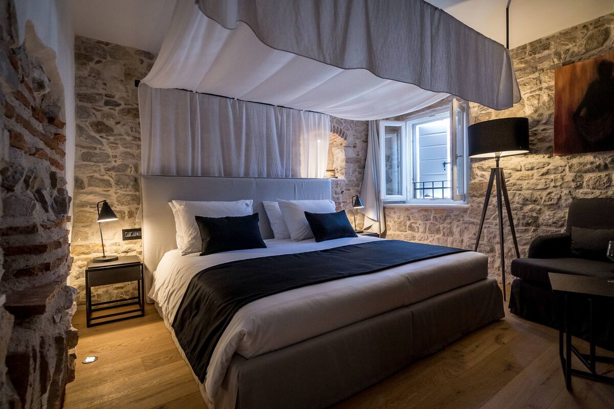 A bedroom with a large bed, a bedside table with a lamp, a chair, and a stone wall.
