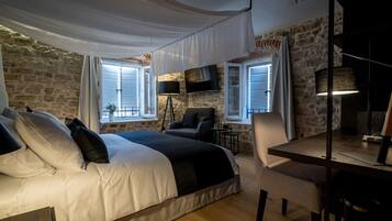 A bedroom with a stone wall, a bed with white and black bedding, a desk with a chair, and a TV mounted on the wall.