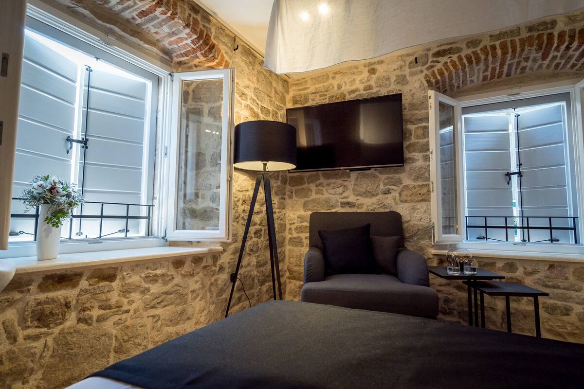 A bedroom with stone walls, a bed, a TV mounted on the wall, and a window with a view.