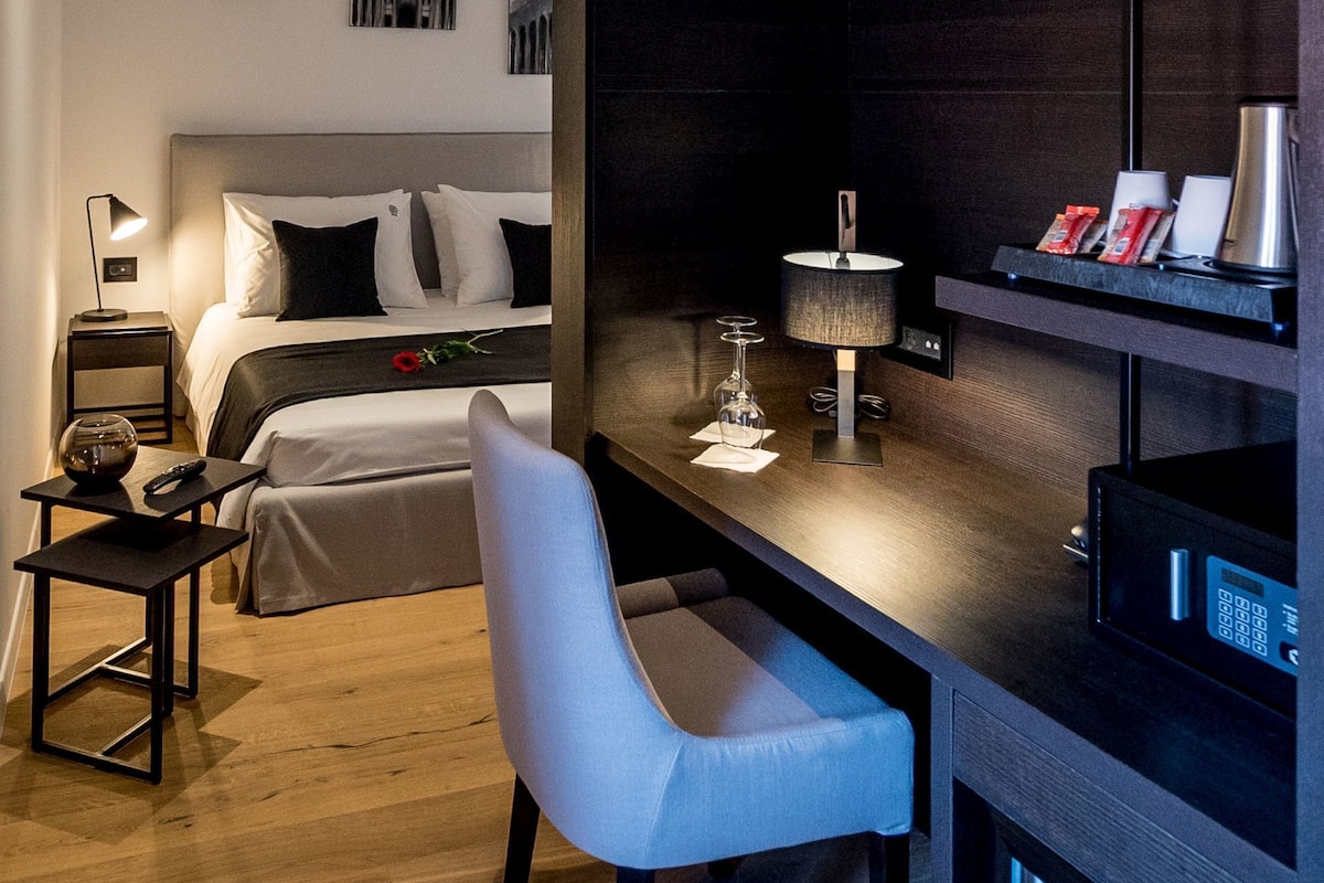A modern hotel room with a bed, bedside table, lamp, chair, and a desk with a computer.
