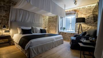Un dormitorio con pared de piedra, una cama con dosel, una mesita de noche con lámpara, un sofá y una pequeña mesa con vasos.