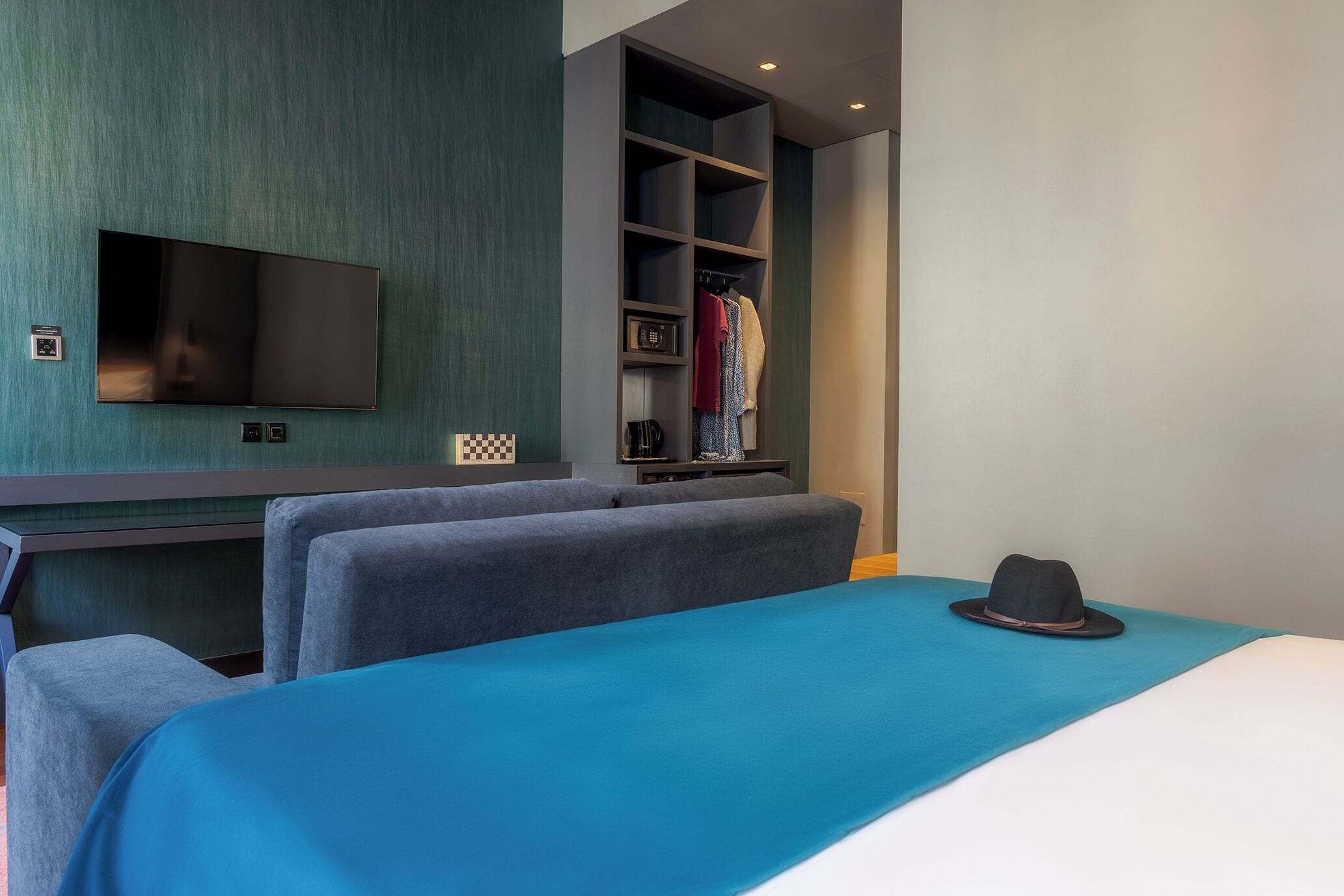 Ein Hotelzimmer mit Bett, Wandfernseher, Kleiderschrank und einem Hut auf dem Bett.