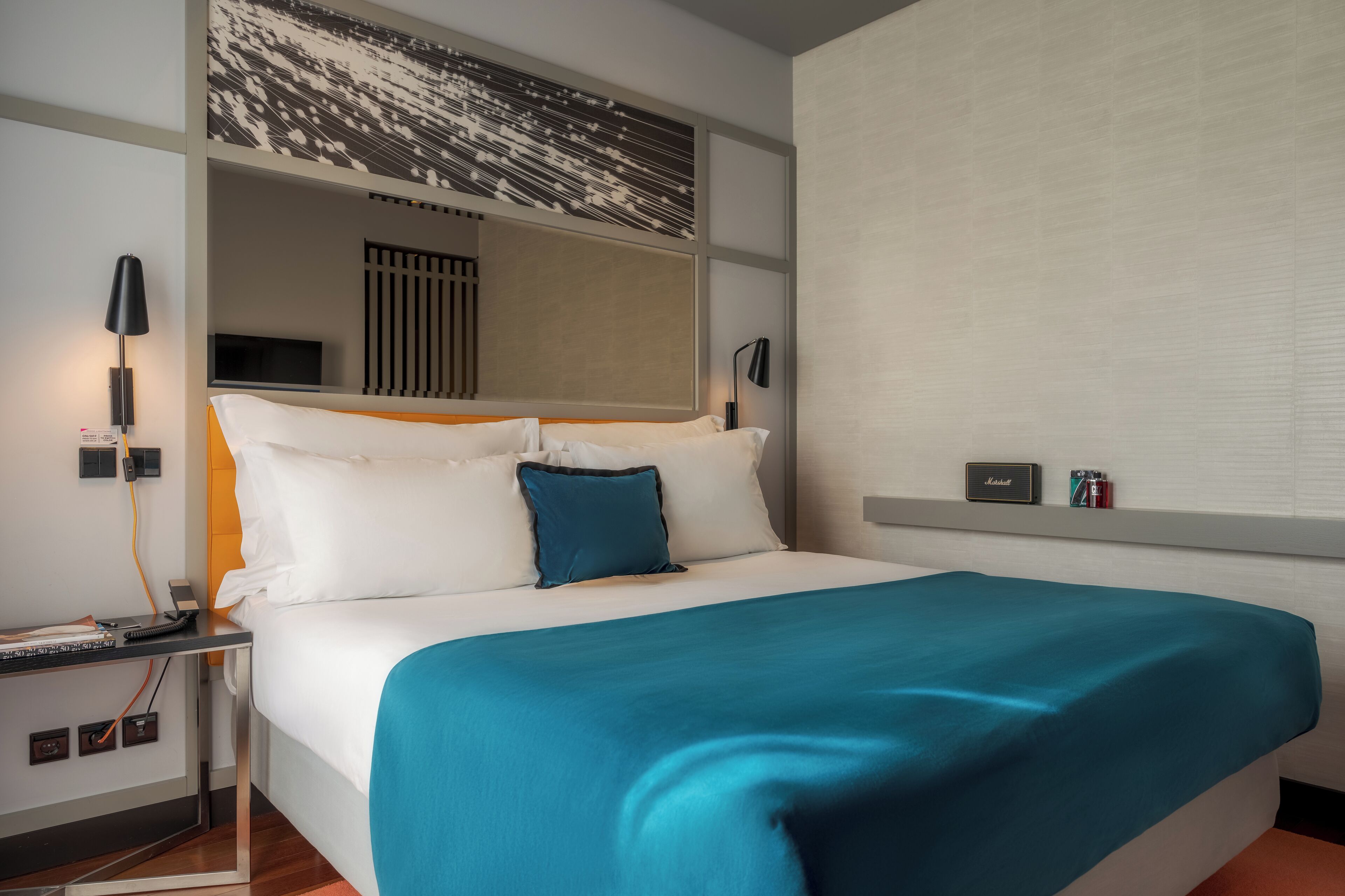 A hotel room with a bed, a nightstand, a lamp, a TV, and a wall-mounted shelf with a nameplate.