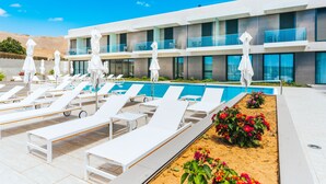 Un hotel moderno con zona de piscina que incluye tumbonas y sombrillas, cielo azul despejado y jardín ajardinado con flores.