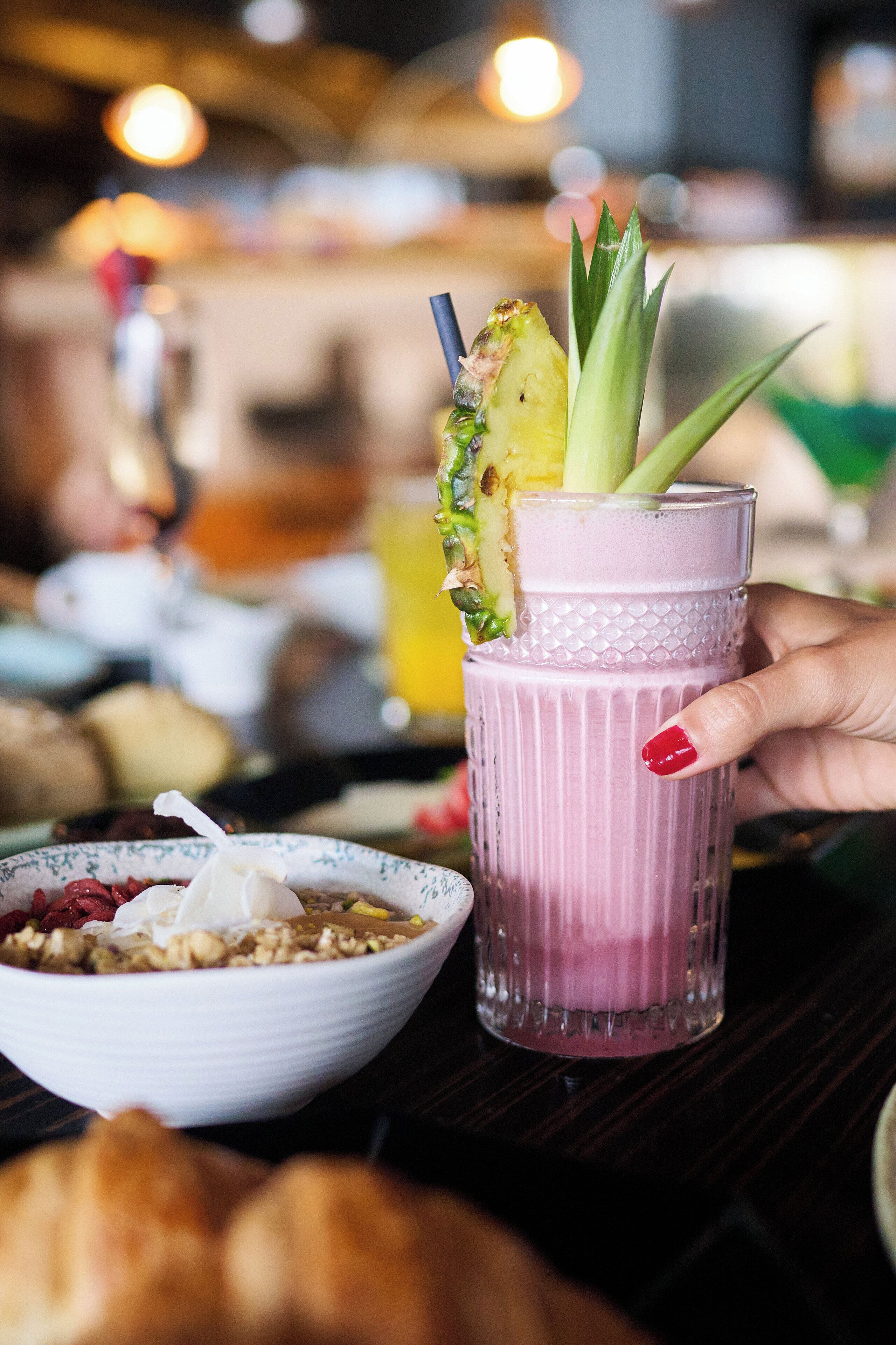 A hand holding a pink beverage with a pineapple and celery garnish, a bowl of granola with yogurt, and a croissant on a table.