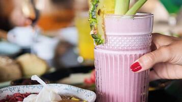 A hand holding a pink beverage with a pineapple and celery garnish, a bowl of granola with yogurt, and a croissant on a table.
