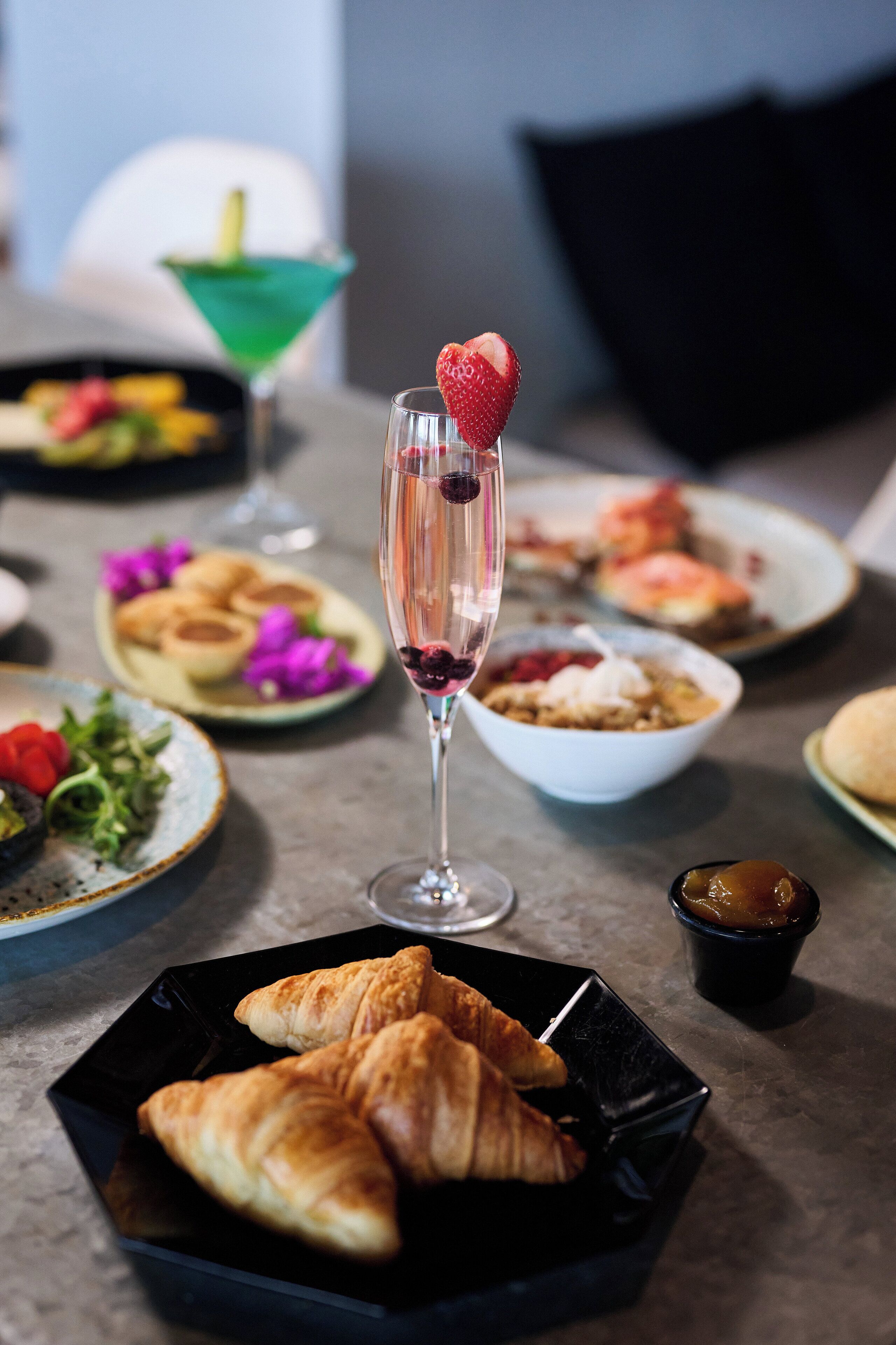 A table with various dishes, including croissants and a glass with a strawberry garnish.