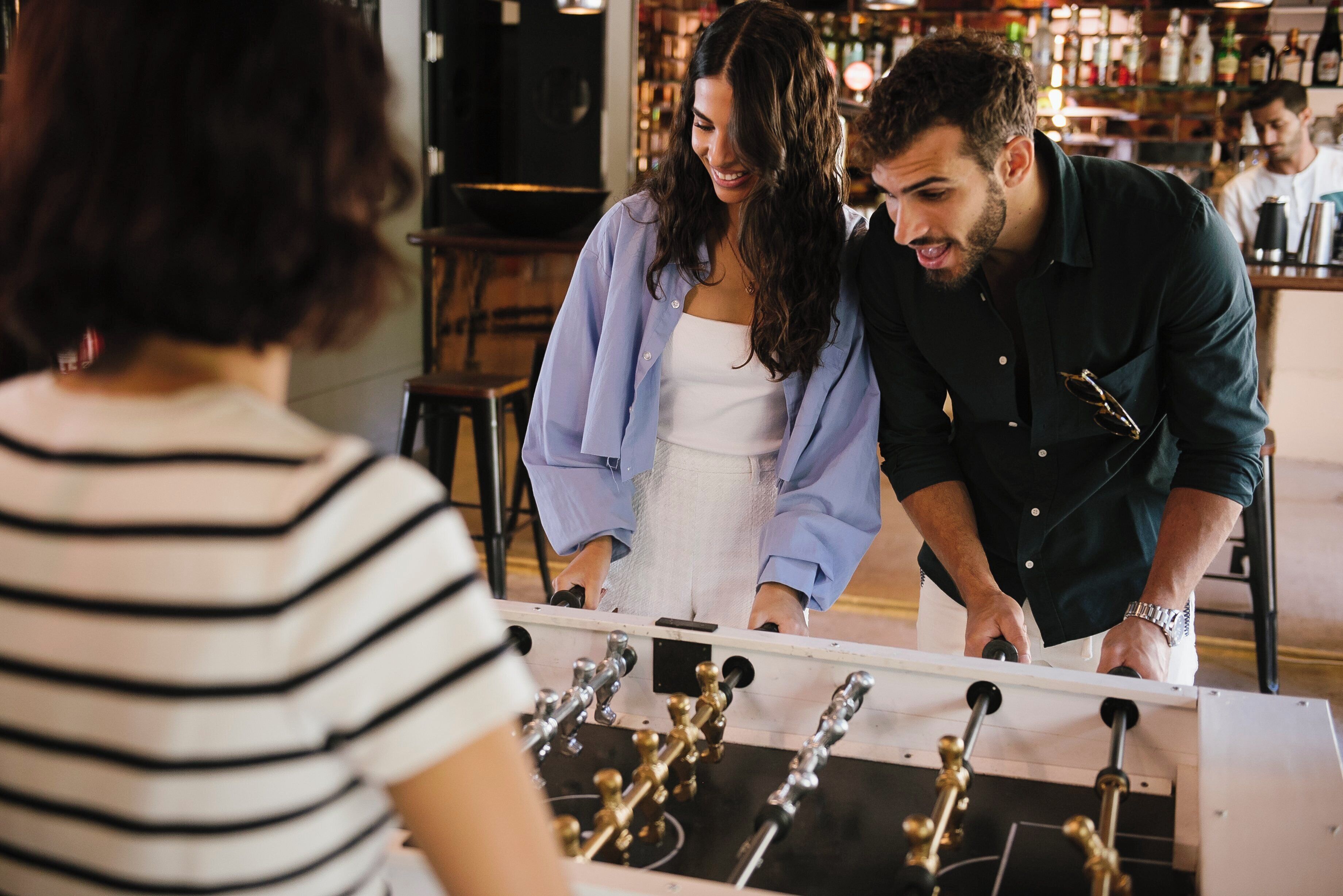 A group of people playing foosball in a bar setting with a bar counter and shelves of bottles in the background.