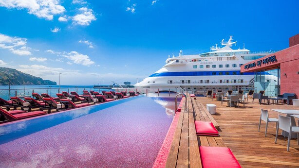 A poolside area with lounge chairs and tables, a large cruise ship in the background, and a building labeled "Asta Nova Cry Hotel". - Pestana CR7 Funchal (Funchal)