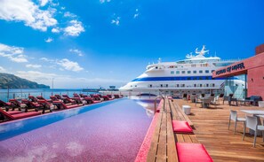 A poolside area with lounge chairs and tables, a large cruise ship in the background, and a building labeled "Asta Nova Cry Hotel". - Pestana CR7 Funchal (Funchal)