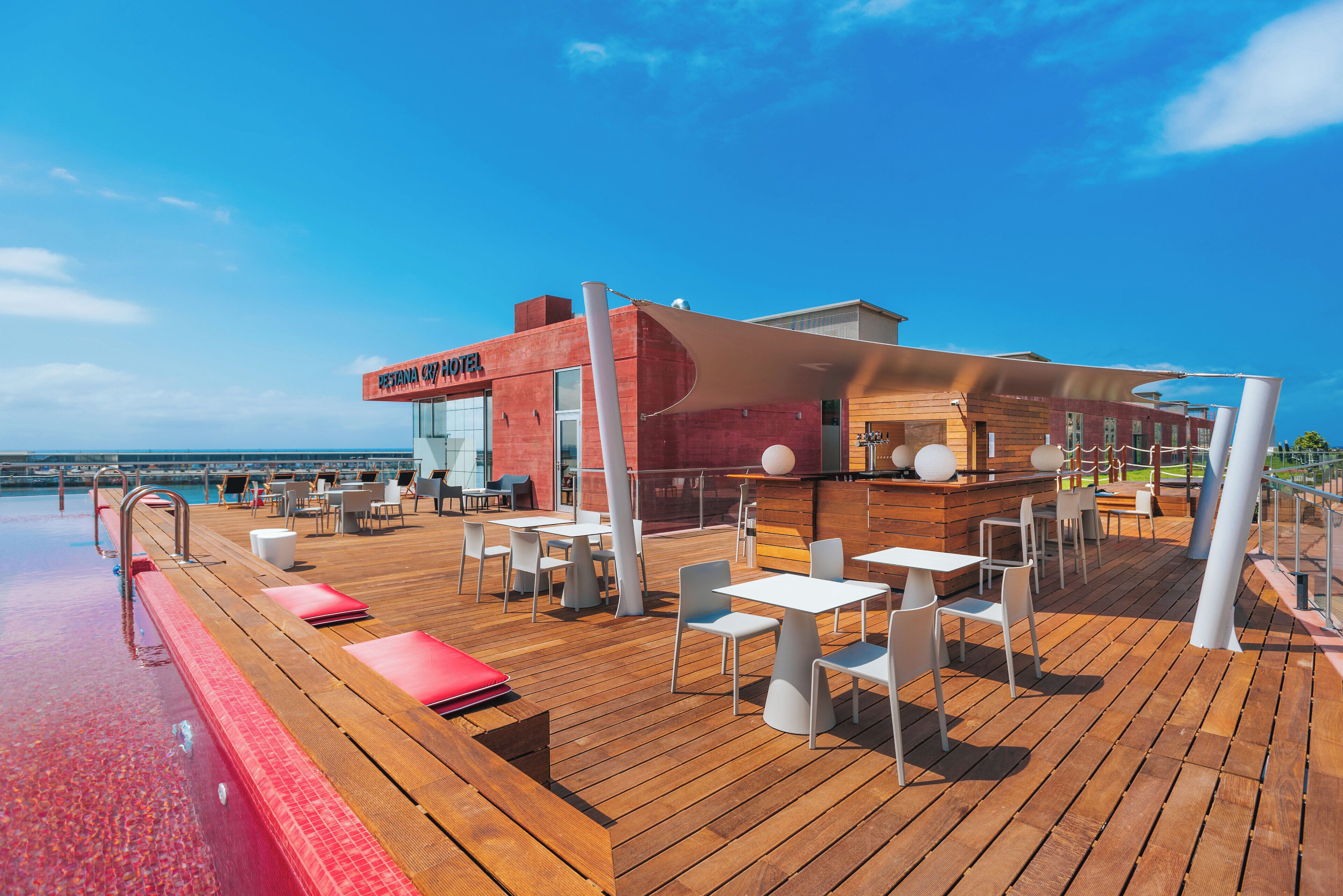 A rooftop bar with wooden deck, white tables, and chairs, overlooking a pool and the ocean.