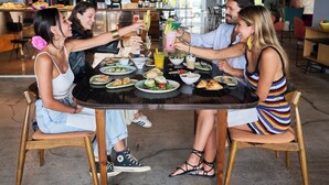 A group of people sitting around a table with various dishes and drinks, in a modern interior with visible kitchen appliances and a bar area in the background. - Pestana CR7 Funchal (Funchal)