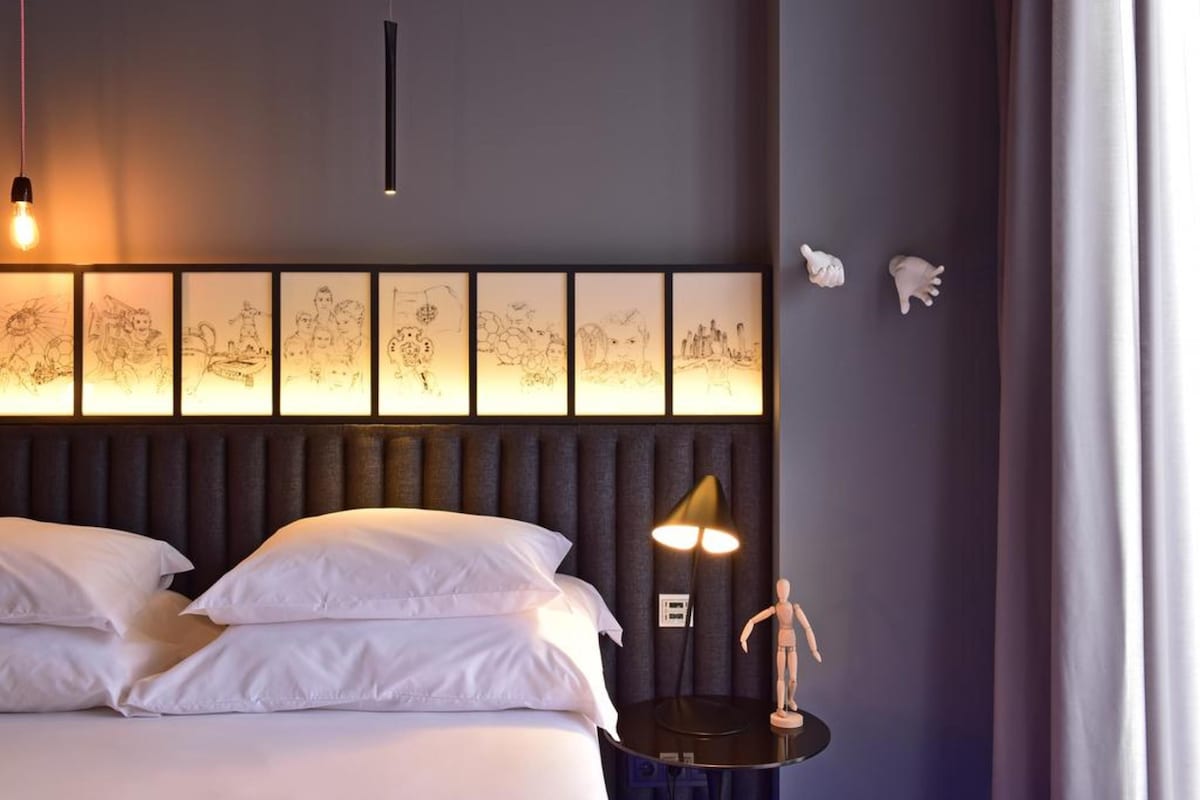 Habitación de hotel con cama, almohadas blancas, mesita de noche con lámpara y una obra de arte en la pared.