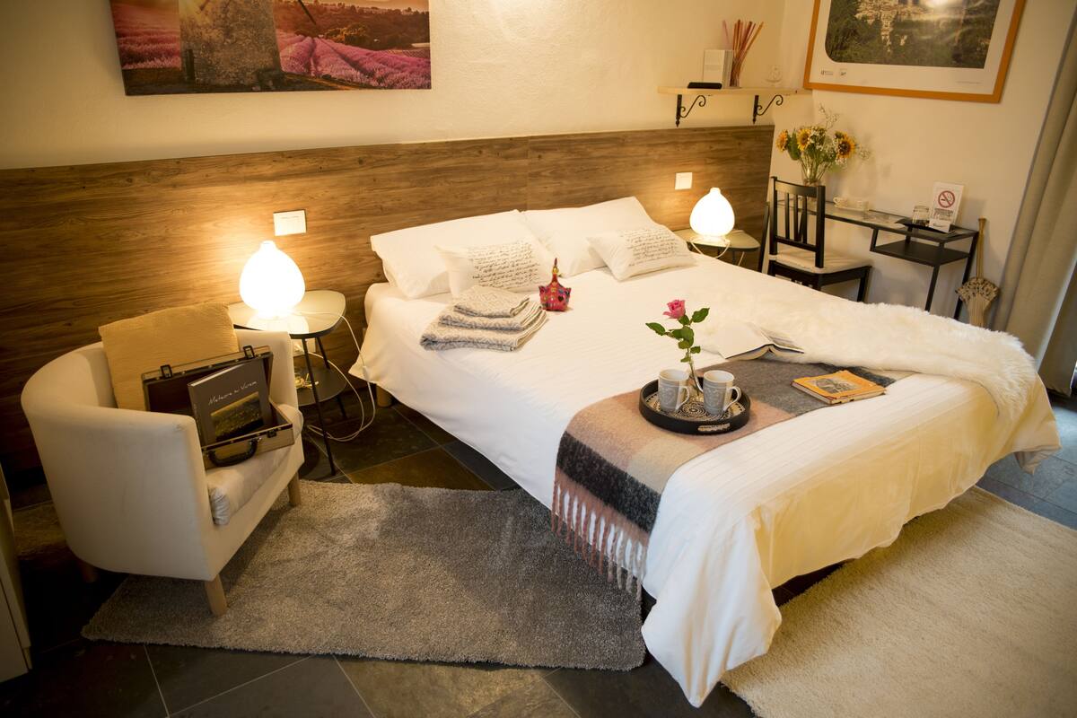 Quarto de hotel bem arrumado, com uma cama grande, um espaço de estar com uma poltrona, uma pequena mesa com abajur e uma prateleira na parede com objetos decorativos.