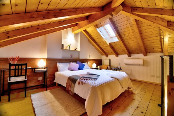 A bedroom with a sloped ceiling, wooden beams, a bed with white bedding, a bedside table with a lamp, and a small shelf with decorative items.