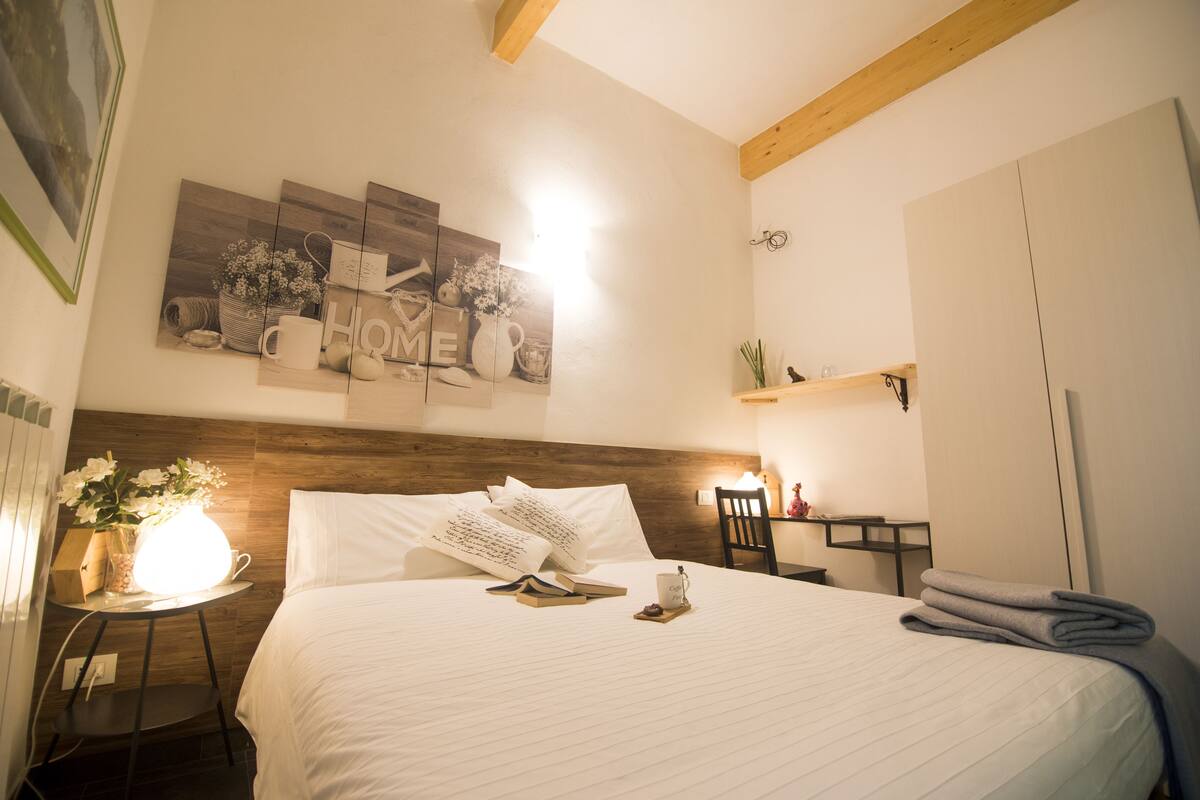 Cama bem arrumada com almofadas e uma xícara, mesa de cabeceira com abajur, e uma parede com um quadro emoldurado.
