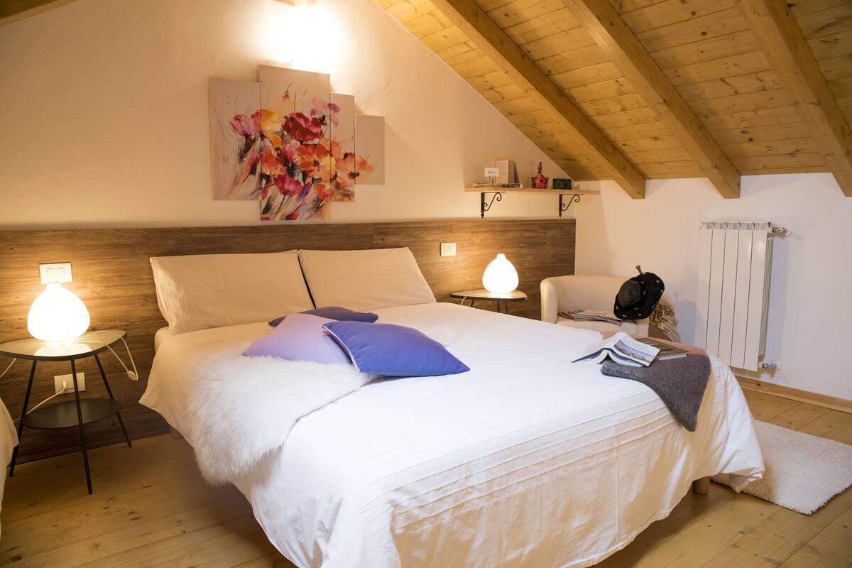 Quarto com teto de madeira, uma cama grande com roupa de cama branca, duas luminárias de cabeceira, uma obra de arte na parede e um radiador.