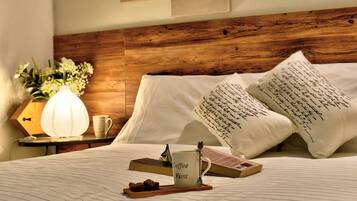 A neatly made bed with a wooden headboard, white pillows with handwritten notes, a bedside table with a lamp, a mug, and a tray with chocolates.