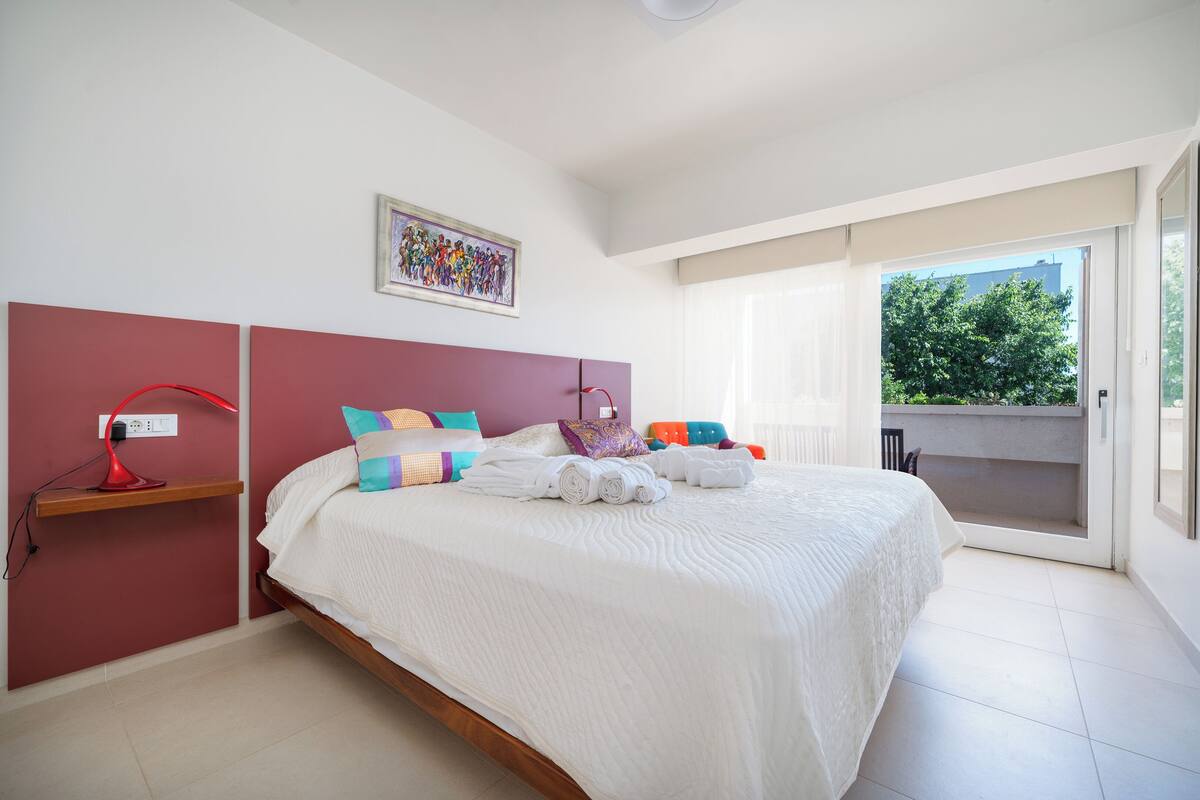 A bedroom with a large bed, a red bedside lamp, a wall-mounted shelf, and a window with a view of greenery.