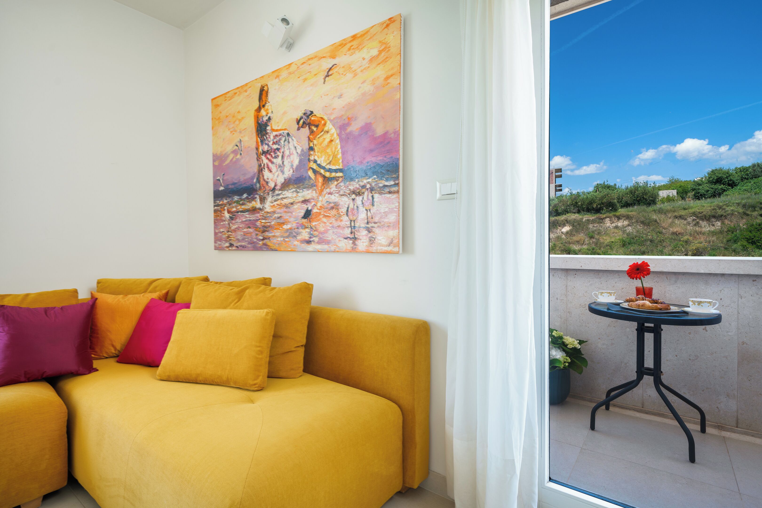 A yellow sofa with multiple cushions, a painting of two figures on a beach, and a balcony with a table and a potted plant.