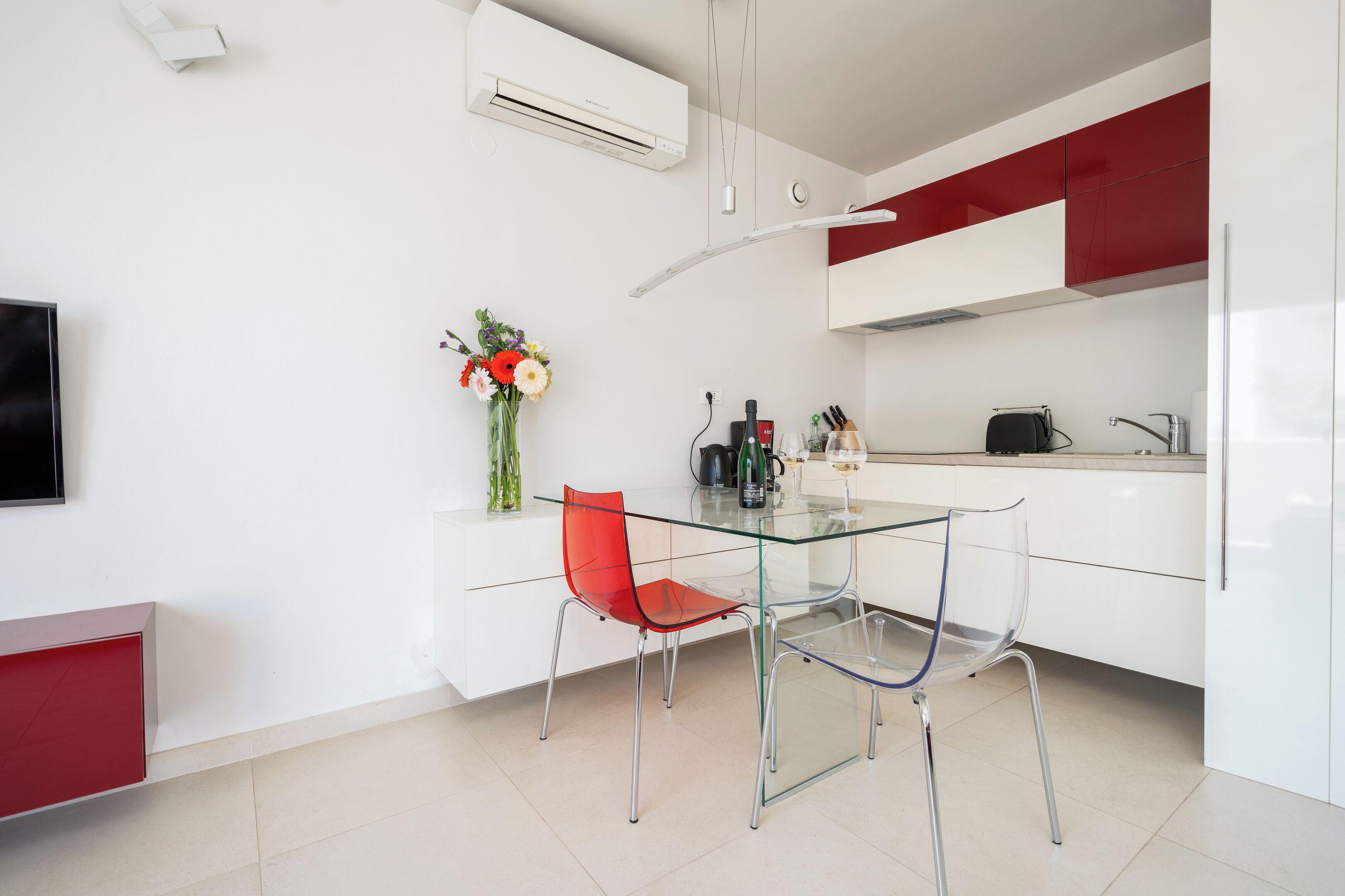 Una cocina moderna con una mesa de comedor de vidrio, sillas rojas, armarios blancos y un florero con flores.