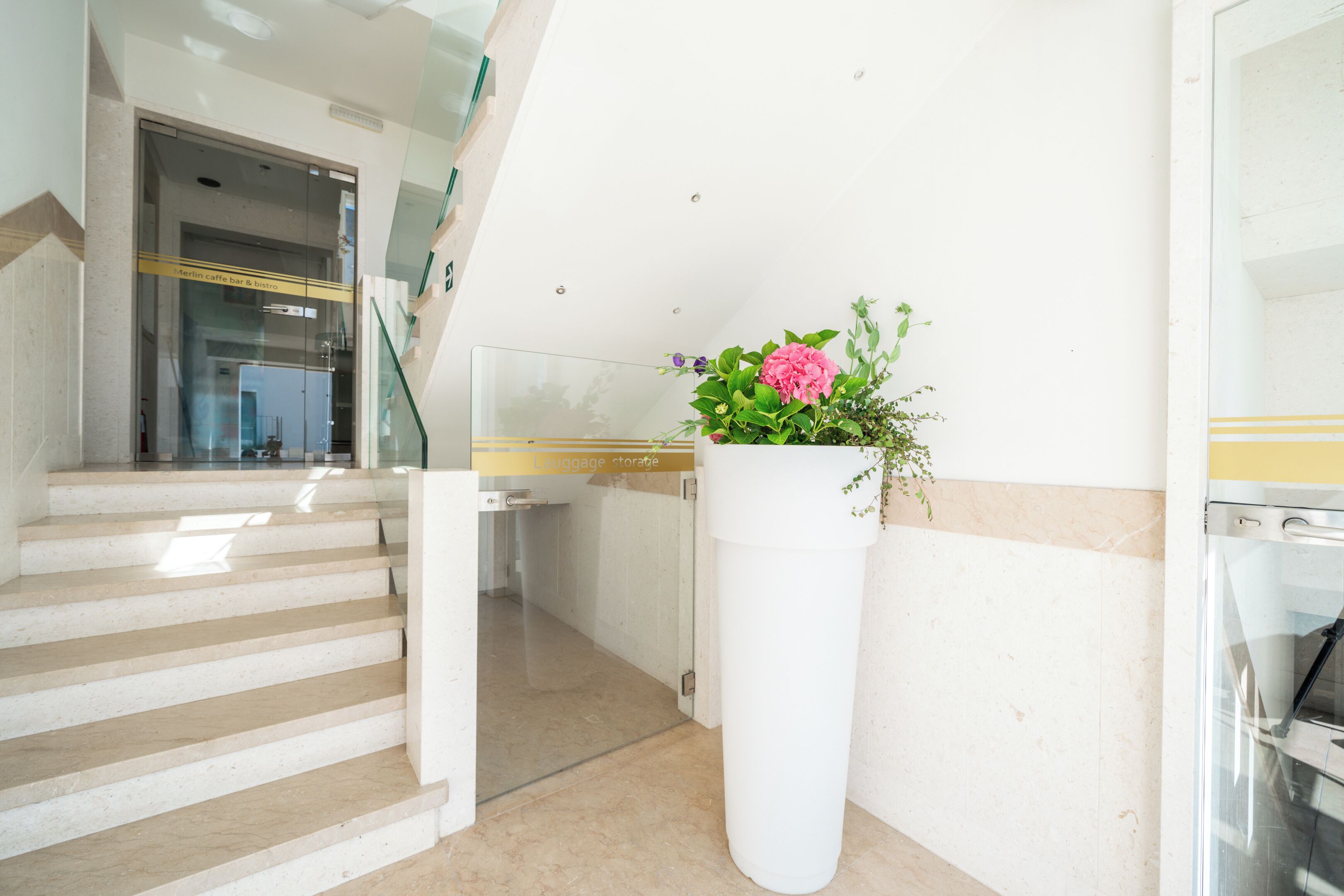 A modern interior with a staircase leading to a lower level, a white column with a flower arrangement, and a visible elevator.