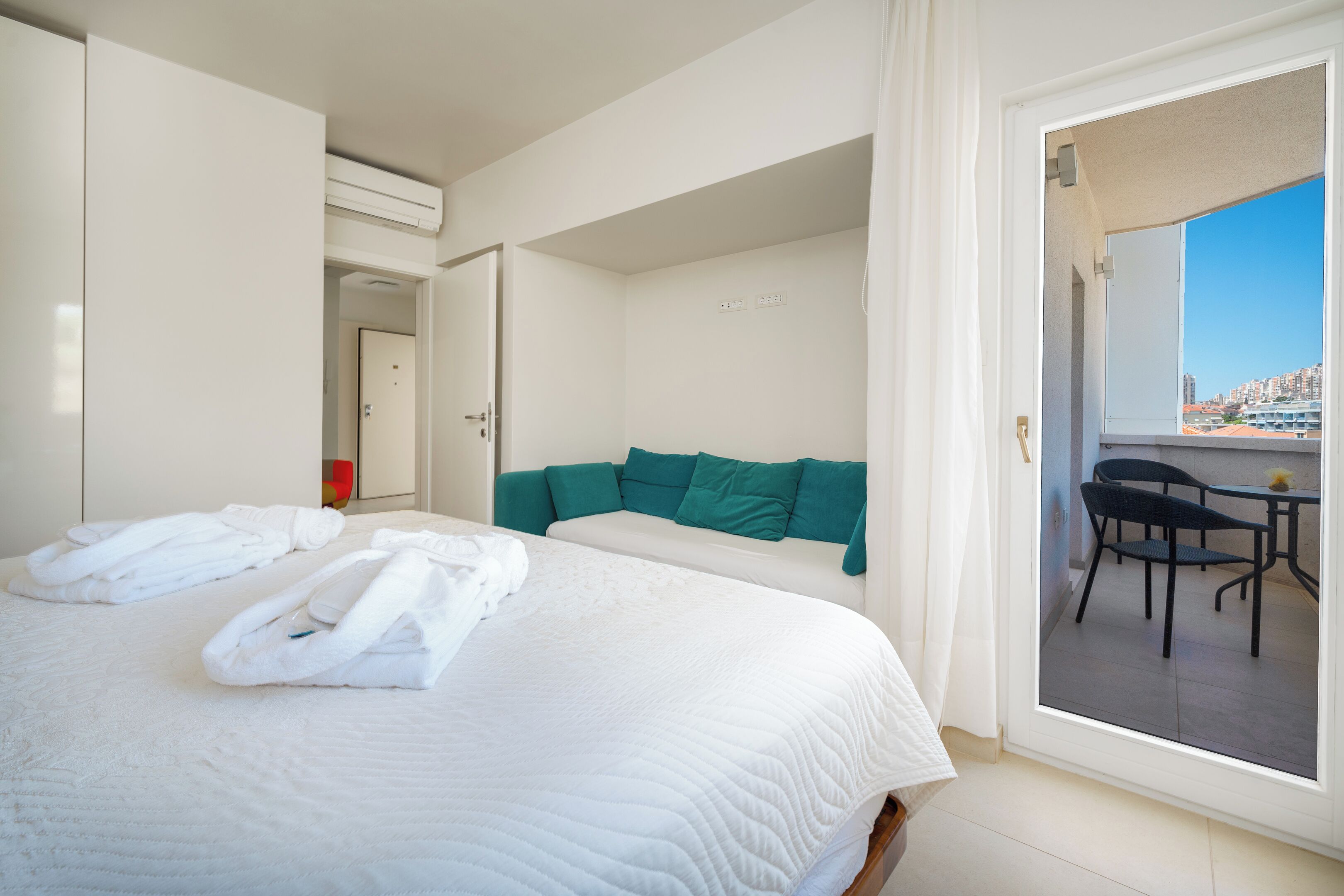 A hotel room with a bed, a green headboard, a balcony with a table and chairs, and a view of the city.