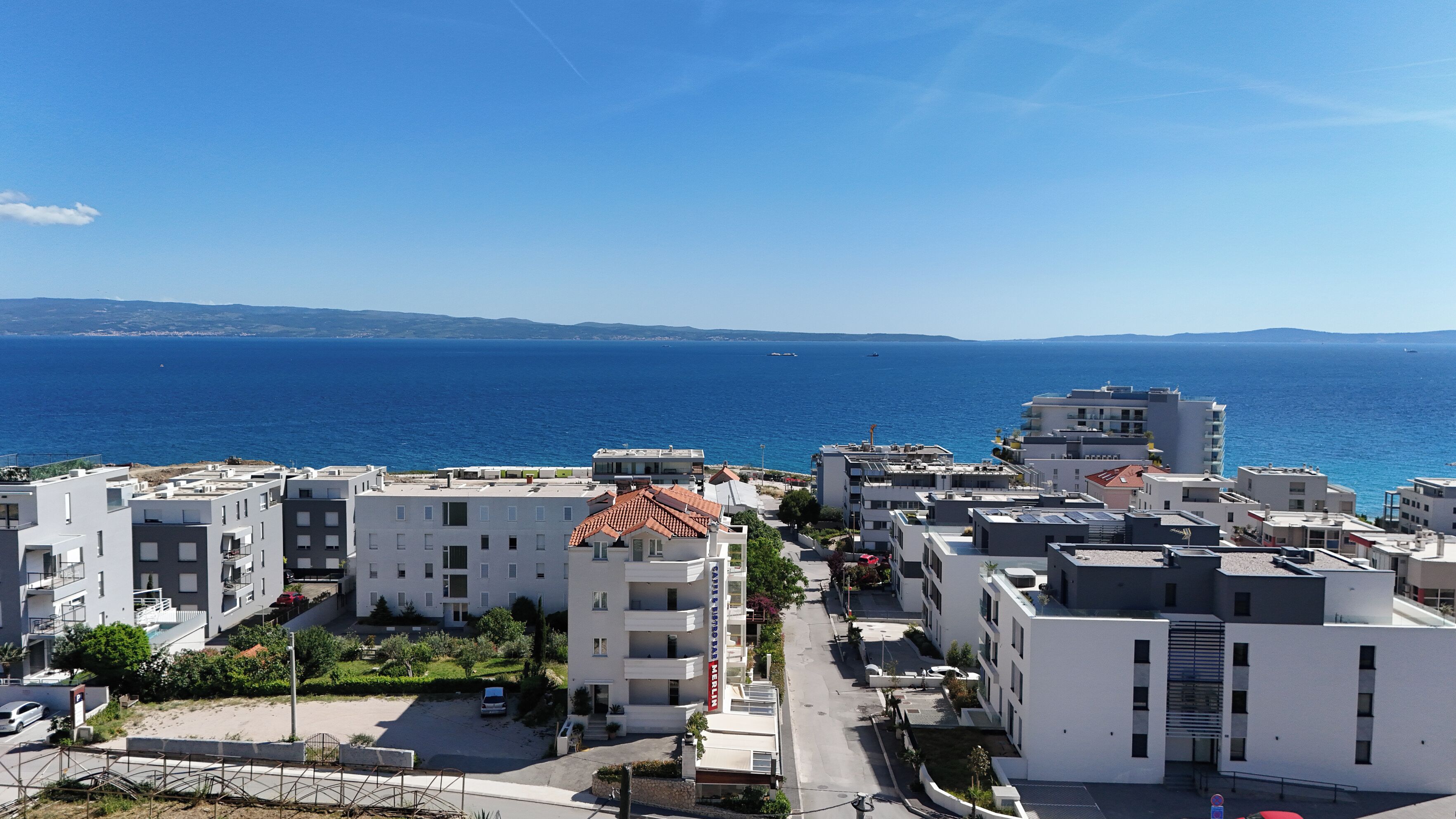 A coastal residential area with modern buildings, a clear blue sky, and the sea in the background.