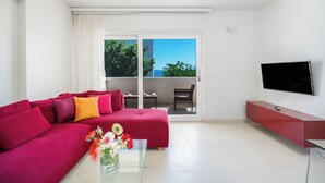 En moderne stue med en rød sofa, et sofabord i glas med blomster, et fladskærms-tv og en skydedør af glas, der fører til en udendørs balkon.