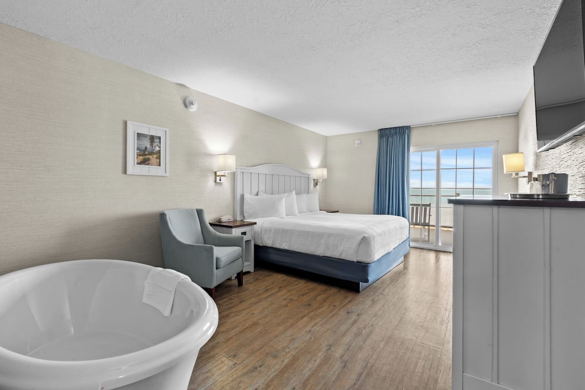 A hotel room with a bed, a chair, a bathtub, and a view of the sea through a large window.