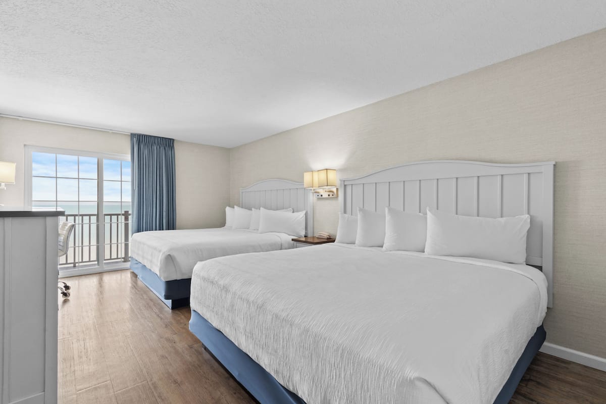 A hotel room with two beds, a balcony with a view, and a desk with a lamp.