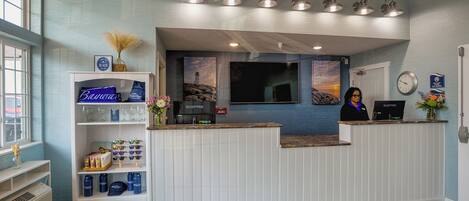 A reception area with a white reception desk, a large flat-screen TV, a clock, and a window with a view of the beach.