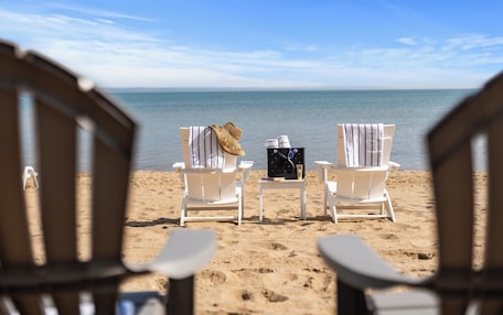 A beach view with two white lounge chairs, one with a straw hat and the other with a towel, and a small table with a book and a bottle between them.