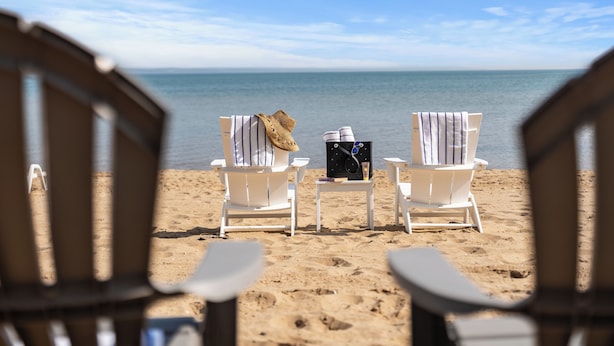 A beach view with two white lounge chairs, one with a straw hat and the other with a towel, and a small table with a book and a bottle between them. - The Baywatch Resort (Traverse City)
