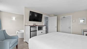 A hotel room with a large bed, a flat-screen TV mounted on the wall, a desk with a chair, a white bathtub, and a door leading to another room.
