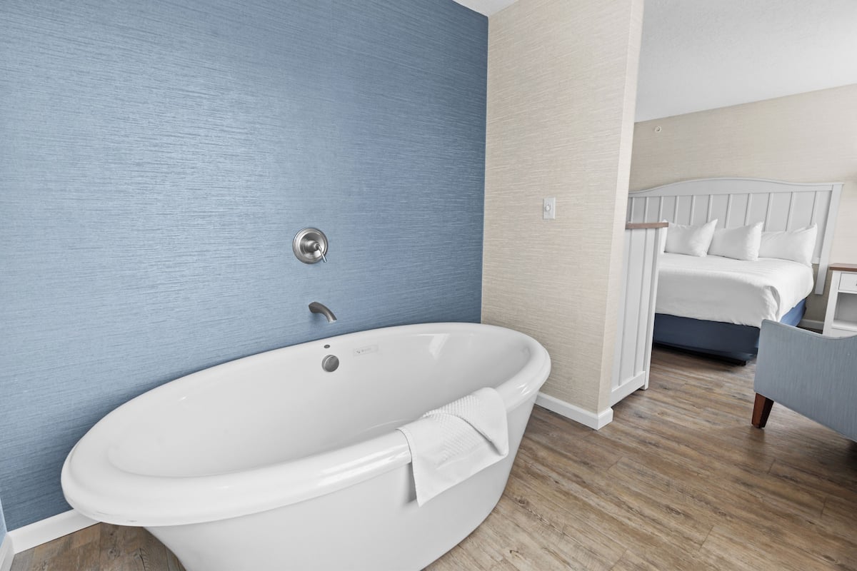 A modern bathroom with a freestanding bathtub, a wooden floor, and a bed in the adjacent room.