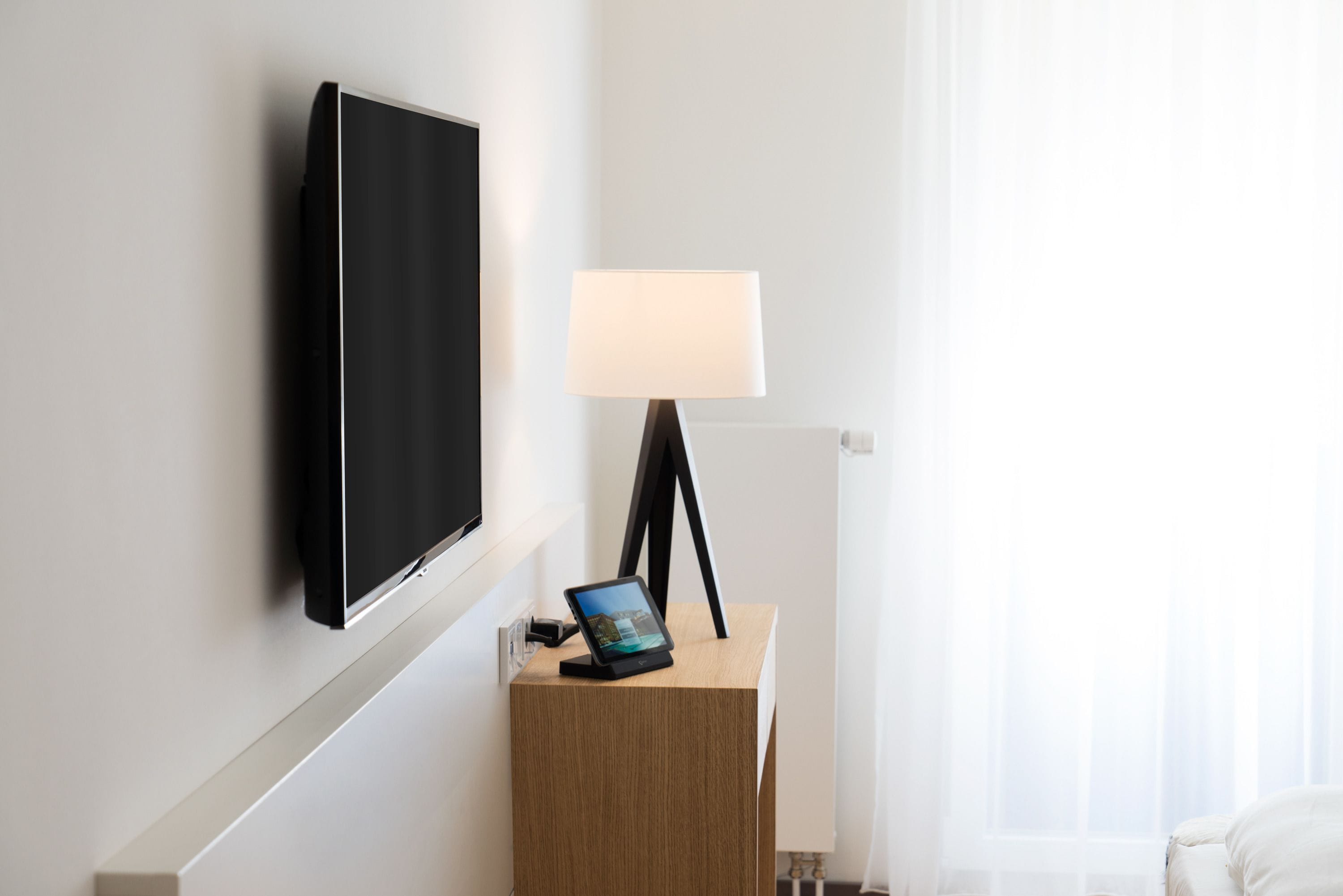 Habitación de hotel con televisión de pantalla plana montada en la pared, una mesita de noche con lámpara, y una tableta sobre la mesa.